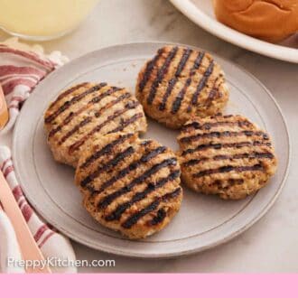 Pinterest graphic of a plate with four chicken burger patties. Burger buns in the background.