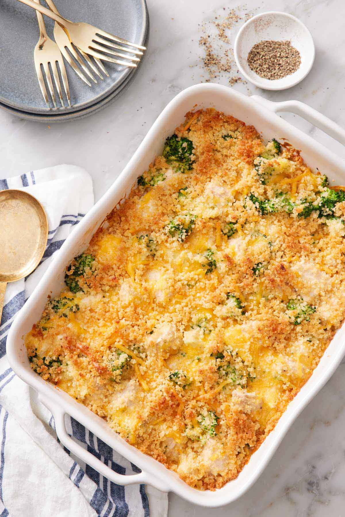 Overhead view of a white baking dish of chicken divan with a bowl of pepper off to the side with a stack of plates and forks.
