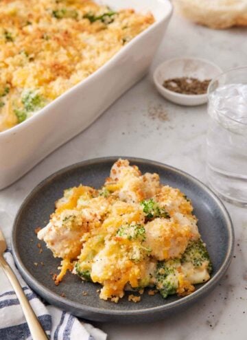A plate with a serving of chicken divan. A baking dish with more in the back along with a glass of water and bowl of pepper.