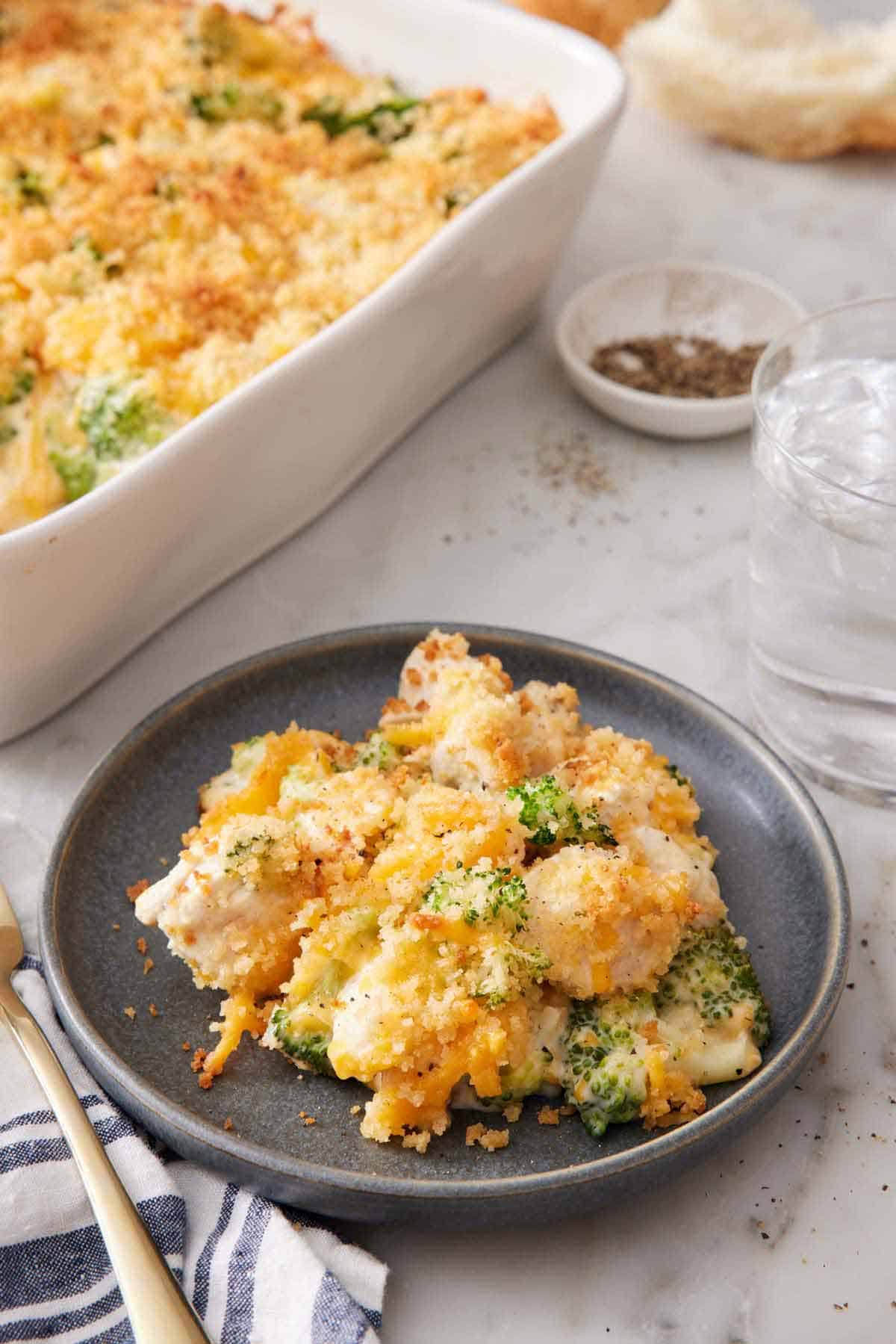 A plate with a serving of chicken divan. A baking dish with more in the back along with a glass of water and bowl of pepper.