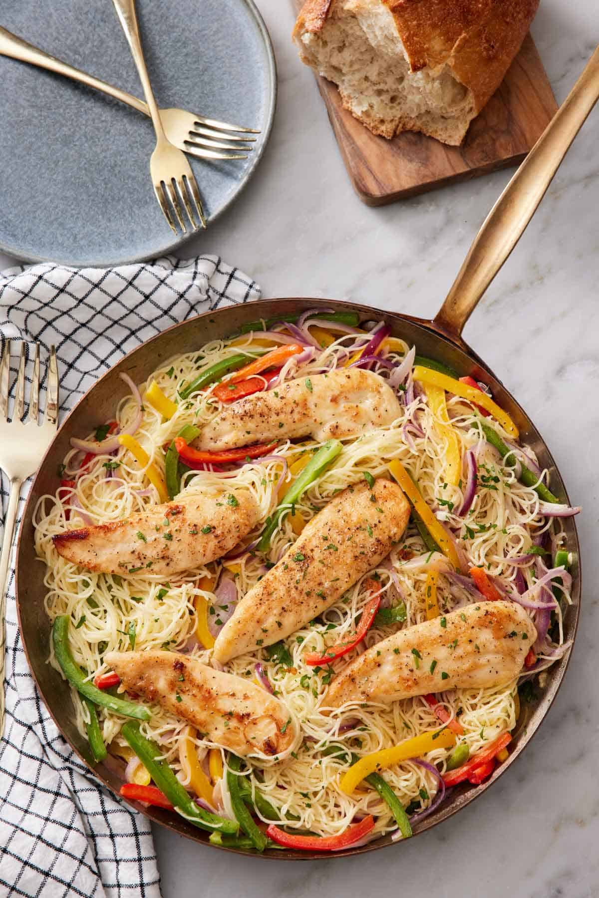 Overhead view of a skillet of chicken scampi with a plate with some forks and a loaf of bread beside it.