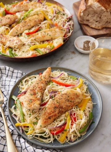 A plate of chicken scampi with a glass of wine, a loaf of bread, and skillet with more chicken scampi in the background.