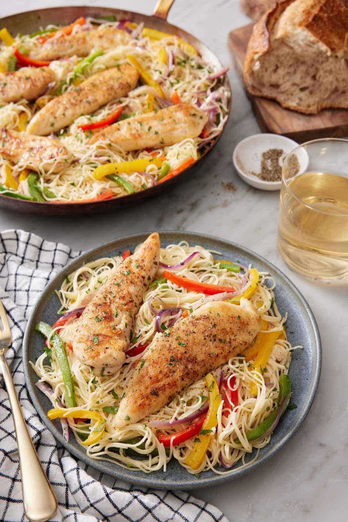 A plate of chicken scampi with a glass of wine, a loaf of bread, and skillet with more chicken scampi in the background.