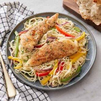 A plate of chicken scampi with two pieces of chicken tenderloin on top. A loaf of bread and a fork on a napkin beside the plate.