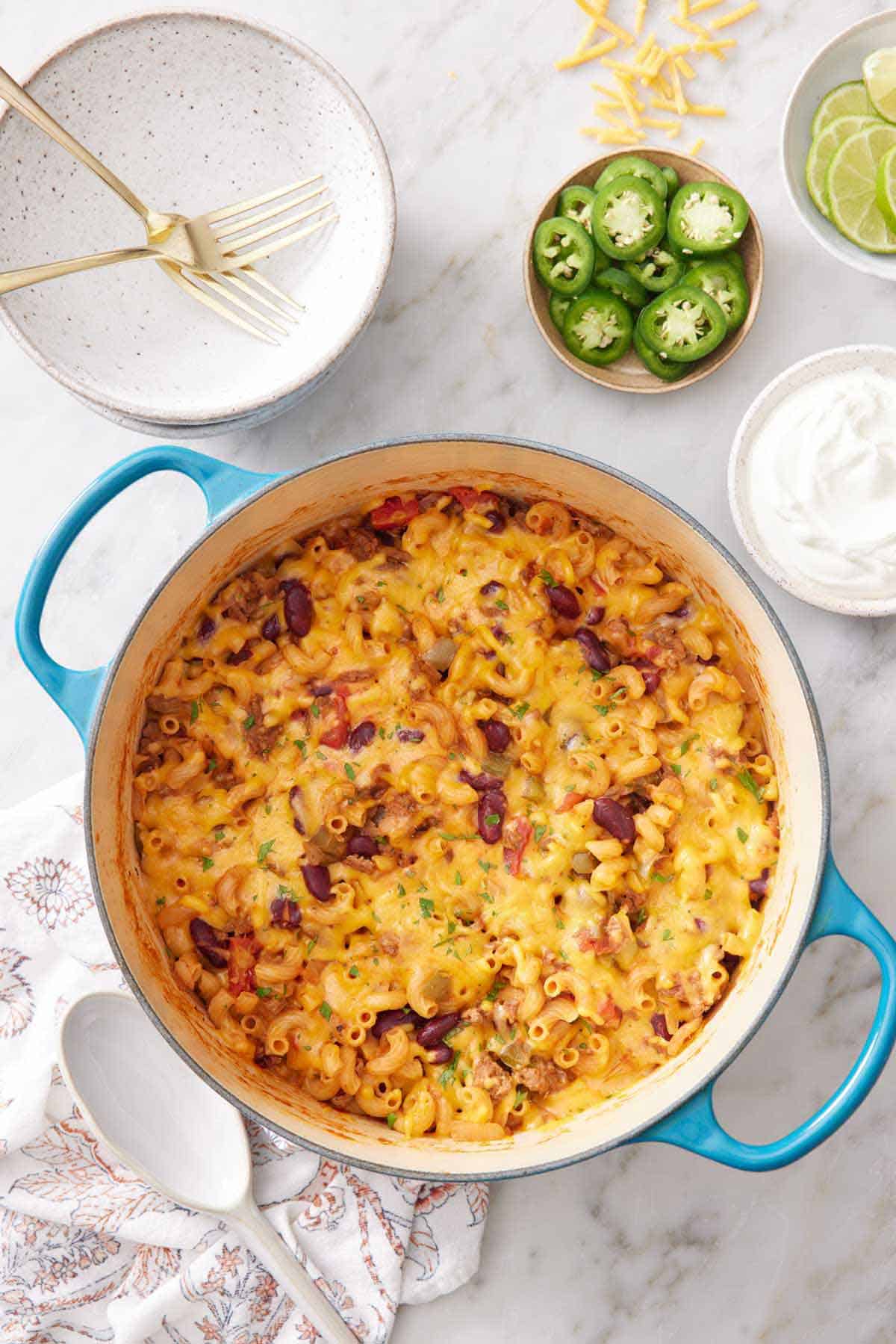 Overhead view of a pot of chili mac with a stack of plates, forks, sliced jalapeño, and sour cream in the background.