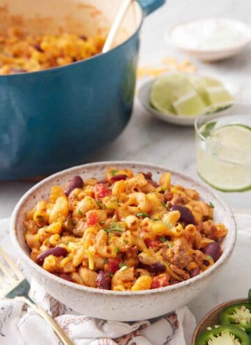 A bowl of chili mac with a drink in the background along with a bowl of limes and a pot of more chili mac.