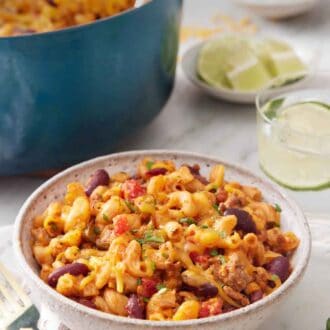 Pinterest graphic of a bowl of chili mac with a drink in the background along with a bowl of limes and a pot of more chili mac.