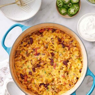 Pinterest graphic of a pot of chili mac with a stack of plates, forks, sliced jalapeño, and sour cream in the background.