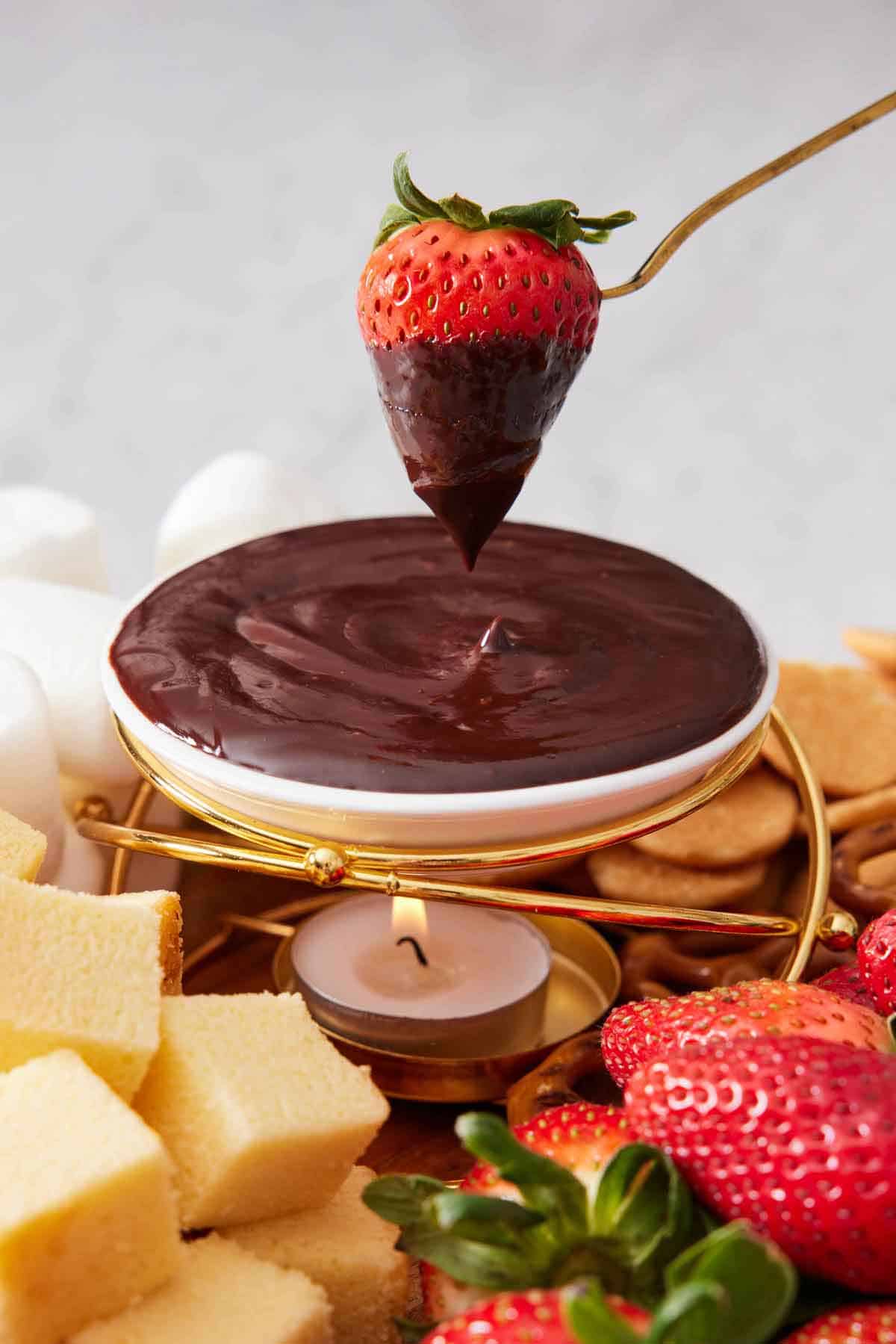 A strawberry coated in chocolate lifted out from a bowl of chocolate fondue, surrounded by more strawberries, cake, and cookies.