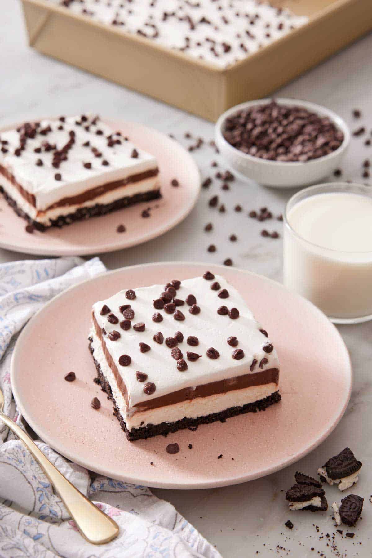 Two plates with sliced chocolate lasagna with a glass of milk, bowl of mini chocolate chips, and a pan of more chocolate lasagna in the background.