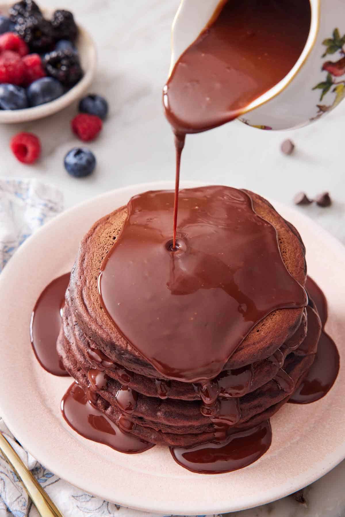 A stack of chocolate pancakes on a plate with chocolate sauce poured on top. Bowl of berries in the background.