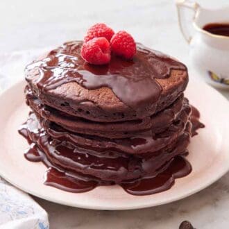 A stack of chocolate pancakes with chocolate sauce and raspberries on top. Berries, chocolate sauce, and a fork on the side.
