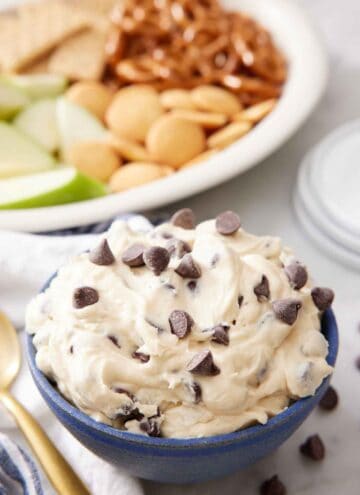 A bowl of cookie dough dip with crackers, pretzels, and apples in the background.