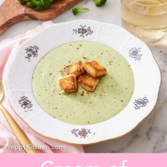 Pinterest graphic of a bowl of cream of broccoli soup topped with croutons and pepper. Broccoli, wine, and croutons in the background.