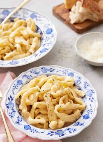 A bowl of cooked egg noodles with a second bowl in the background along with a bowl of parmesan and torn bread.