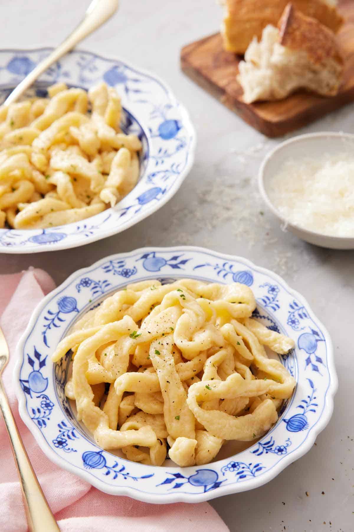 A bowl of cooked egg noodles with a second bowl in the background along with a bowl of parmesan and torn bread.