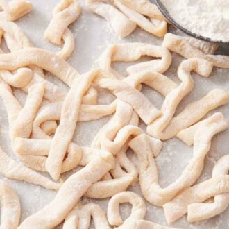 Multiple pieces of uncooked egg noodles coated in flour on a marble surface with a bowl of flour off to the side.