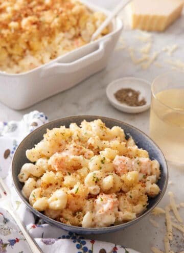 A bowl of lobster mac and cheese with a baking dish with more in the background along with a drink and bowl of pepper.
