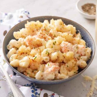 A bowl of lobster mac and cheese with a drink in the background along with a bowl of pepper with a napkin and fork beside it.