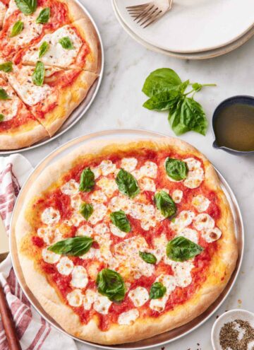 A margherita pizza topped with fresh basil leaves with a second sliced pizza in the background along with a stack of plates and forks.
