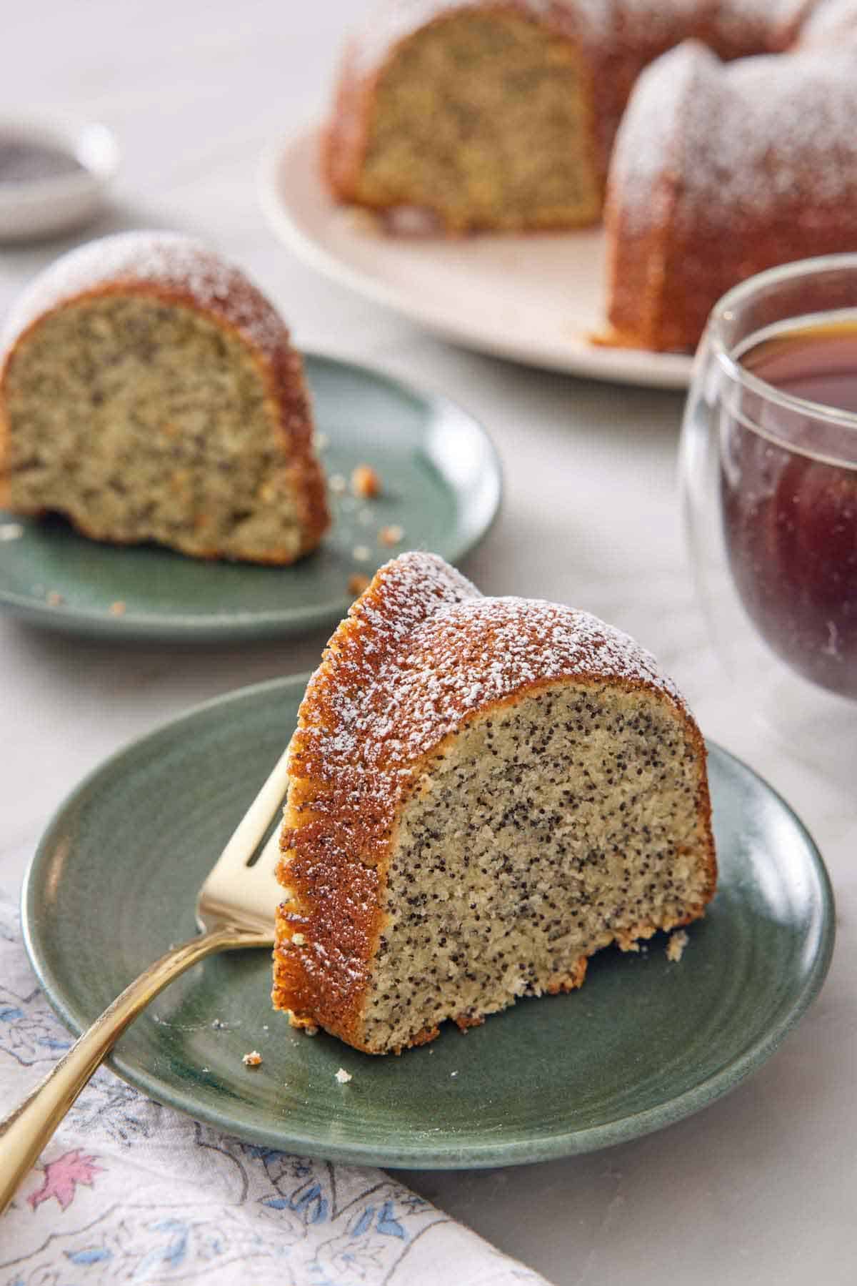 A slice of poppy seed cake on a plate with a fork. Another plated slice in the background along with the rest of the cake.