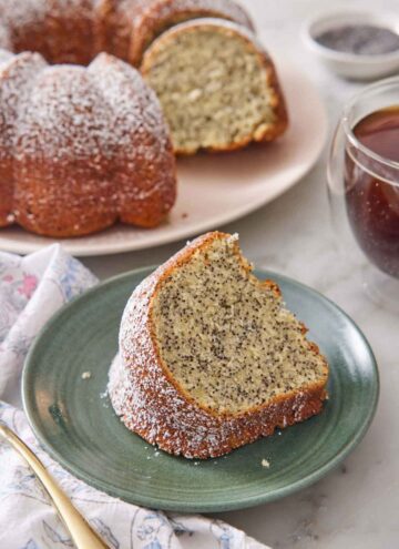 A slice of poppy seed cake on a green plate with the rest of the cake in the background.