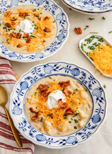 Two bowls of potato soup with shredded cheese, bacon, sour cream, and chives on top.