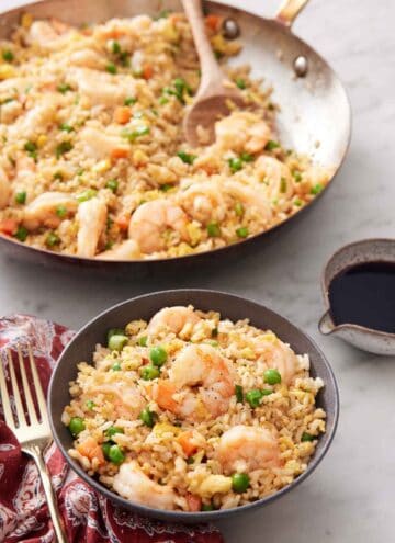 A bowl of shrimp fried rice with a skillet with more in the background with a wooden spoon.