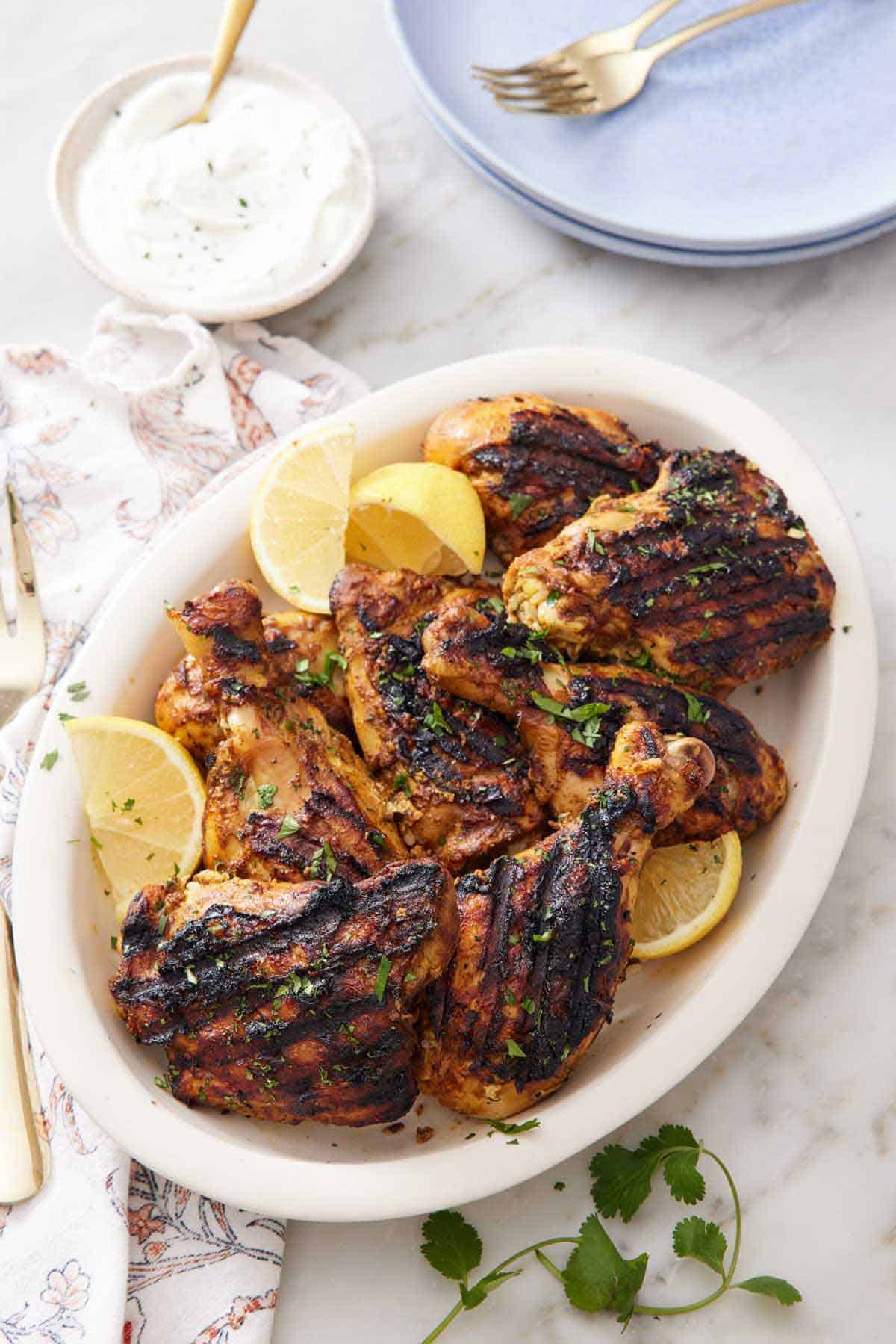 A platter of tandoori chicken topped with chopped parsley with lemon wedges. Yogurt sauce in the back with a stack of plates.