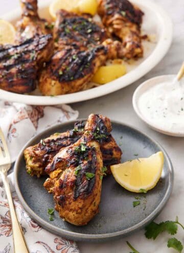 A plate with two pieces of tandoori chicken with a lemon wedge with a yogurt sauce in the background with the rest of the chicken on a platter.