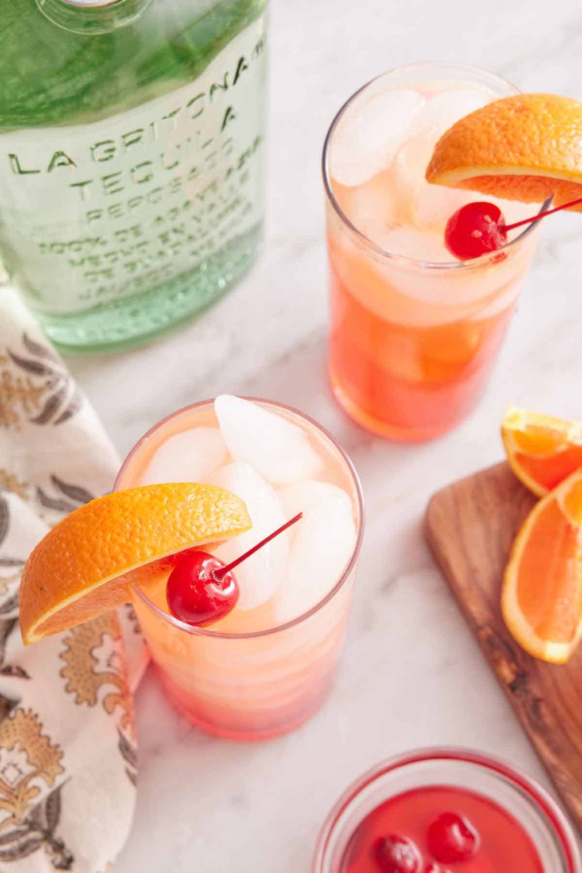 Overhead view of two glasses of tequila sunrises with orange slices and maraschino cherries as garnish. A bottle of tequila beside it.