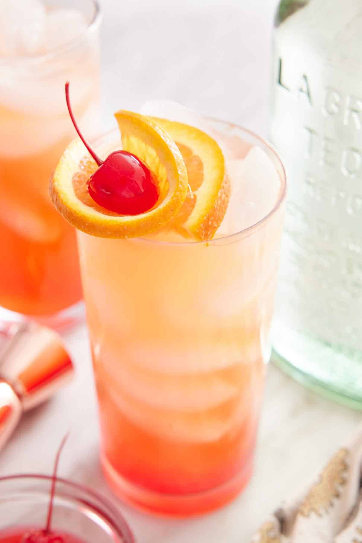 A glass of tequila sunrise with orange slice and maraschino cherry garnish.