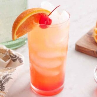 A glass of tequila sunrise cocktail with orange slice and maraschino cherry garnishes on top with ice. More garnishes off to the side.