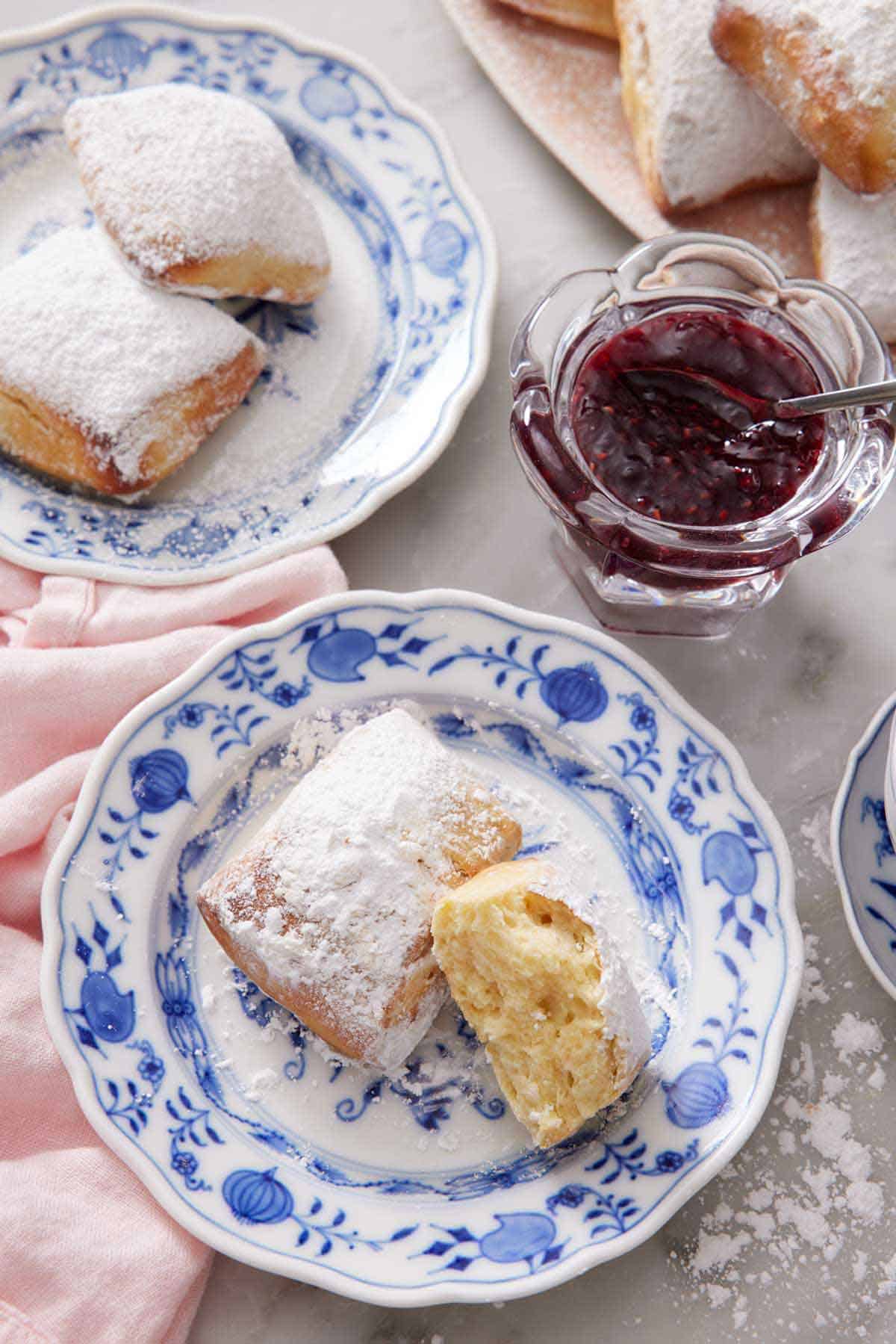 Overhead view of a plate with a whole air fryer beignets and one half, showing the interior. Jar of jam and a plate of two more air fryer beignets behind it.