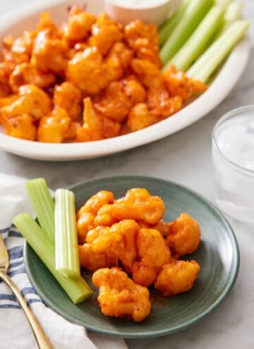A plate with air fryer buffalo cauliflower and celery sticks with more on a platter in the background.