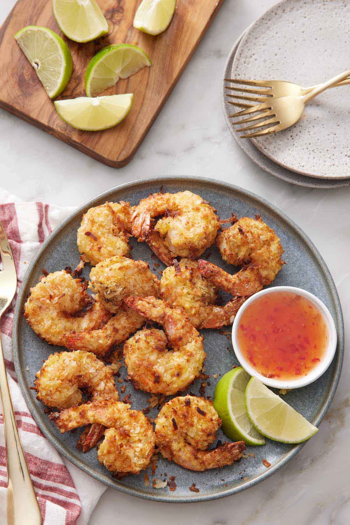 Overhead view of a plate of air fryer coconut shrimp with lime wedges and a bowl of sweet chili sauce. A plate with forks and lime wedges off to the side.