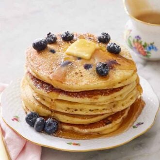 A stack of blueberry pancakes with blueberries, butter, and maple syrup on top with a dusting of powdered sugar.