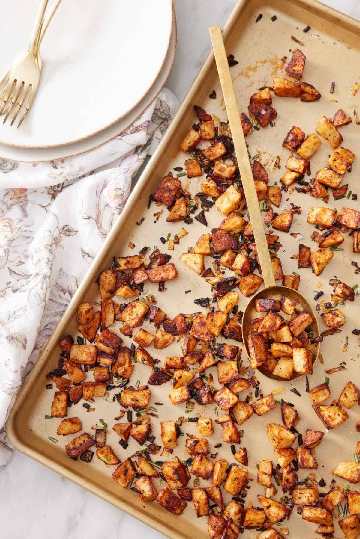 An overhead view of a sheet pan of breakfast potatoes with a ladle with a scoop of potatoes in the middle. A stack of plates, forks, and a napkin on the side.