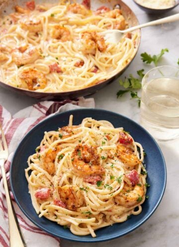 A plate of cajun shrimp pasta with chopped parsley garnish with a skillet with more pasta in the background. A glass of win on the side.