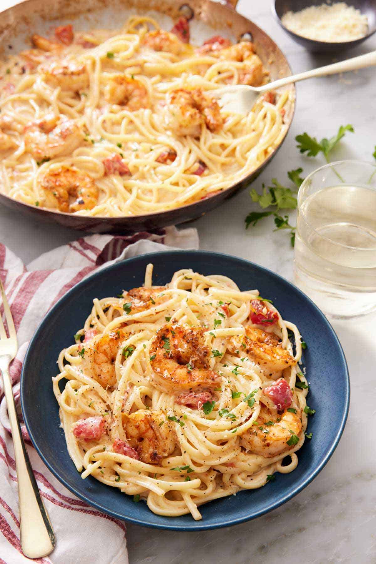 A plate of cajun shrimp pasta with chopped parsley garnish with a skillet with more pasta in the background. A glass of win on the side.