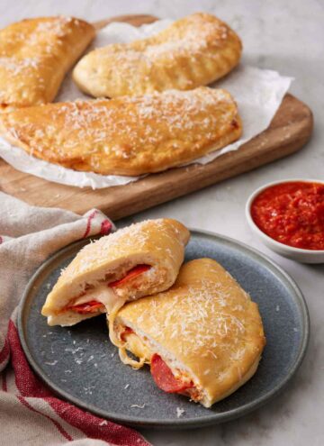 A plate with a calzone cut in half, showing the interior. More calzones in the background along with a bowl of sauce.