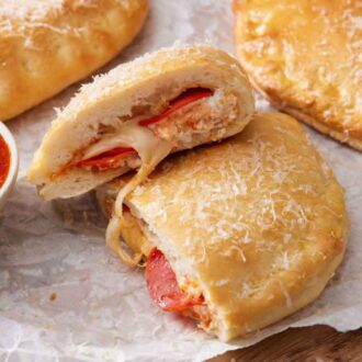 A calzone cut in half with one half stacked on top of the other. Two more in the background with a bowl of pizza sauce on the side.