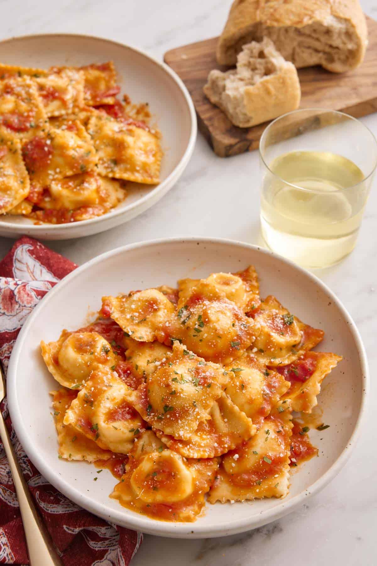 A plate of cheese ravioli tossed in tomato sauce with a glass of wine, torn bread, and another plate of ravioli in the background.