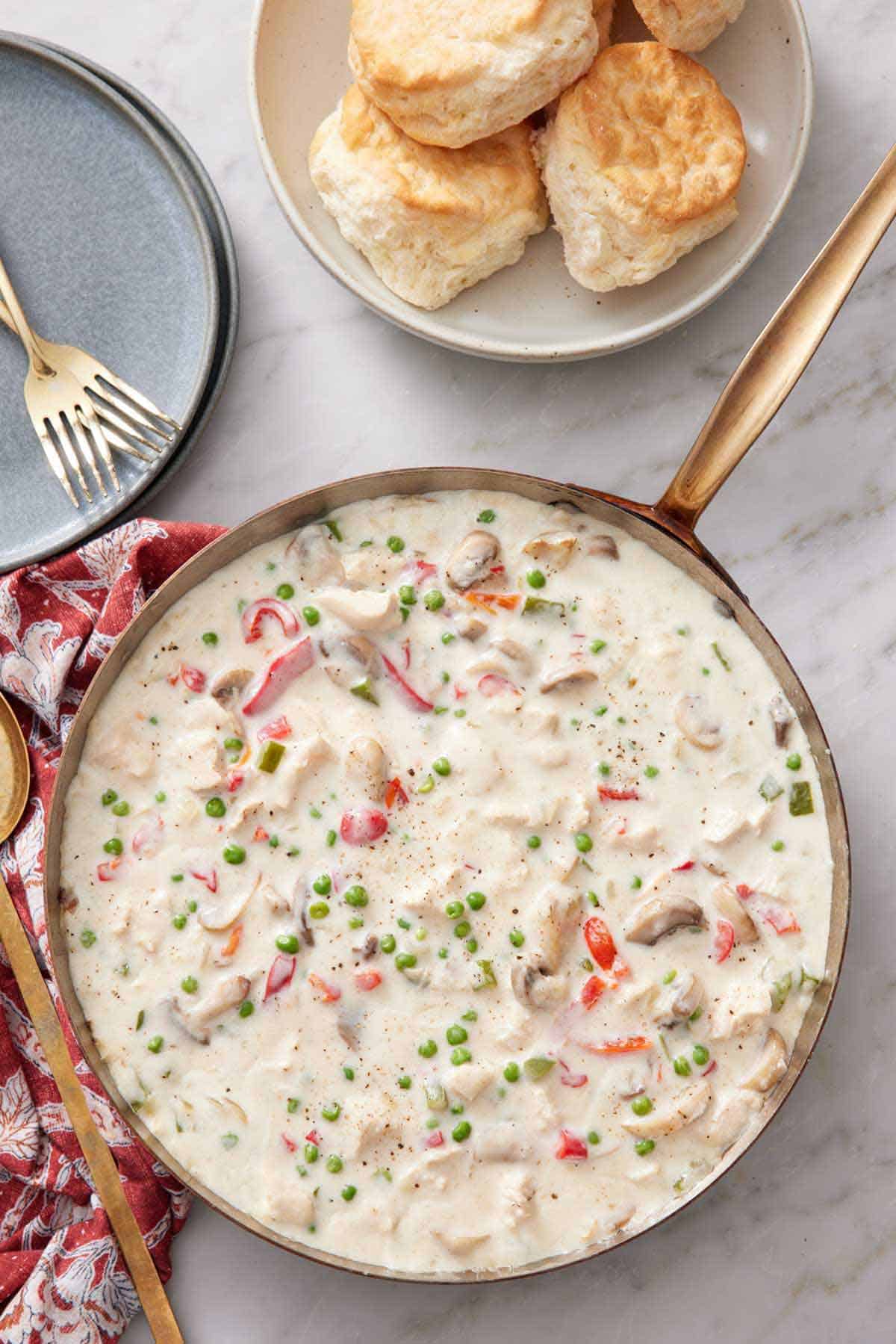 Overhead view of a skillet of chicken a la king with a plate of biscuits on the side along with a stack of plates and forks.