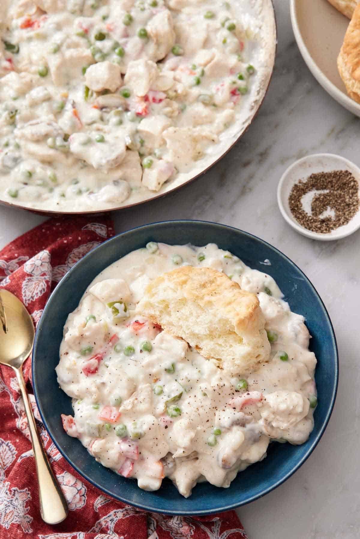 Overhead view of a bowl of chicken a la king with half a biscuit on top. A skillet with more along with a bowl of pepper on the side.