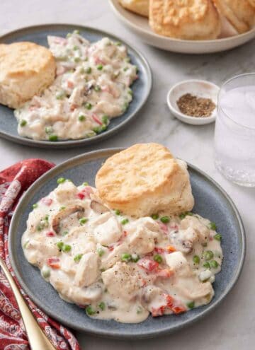 Two plates of chicken a la king with biscuits. A glass of water, a bowl of pepper, and bowl of more biscuits off to the side.