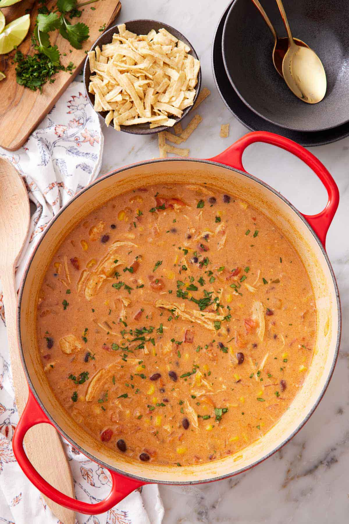 Overhead view of a pot of chicken enchilada soup with a bowl of tortilla strips on the side along with chopped cilantro, limes, and a bowl with spoons.