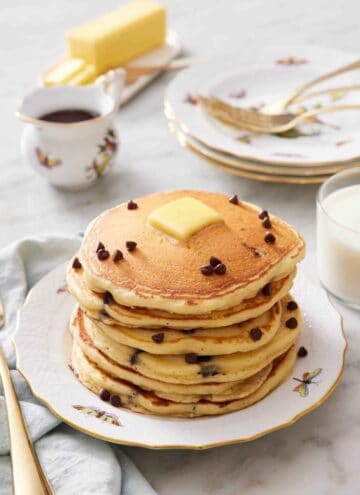 A stack of chocolate chip pancakes with a pat of butter on top with a few extra chocolate chips. A stack of plates and forks in the background along with syrup, butter, and milk.