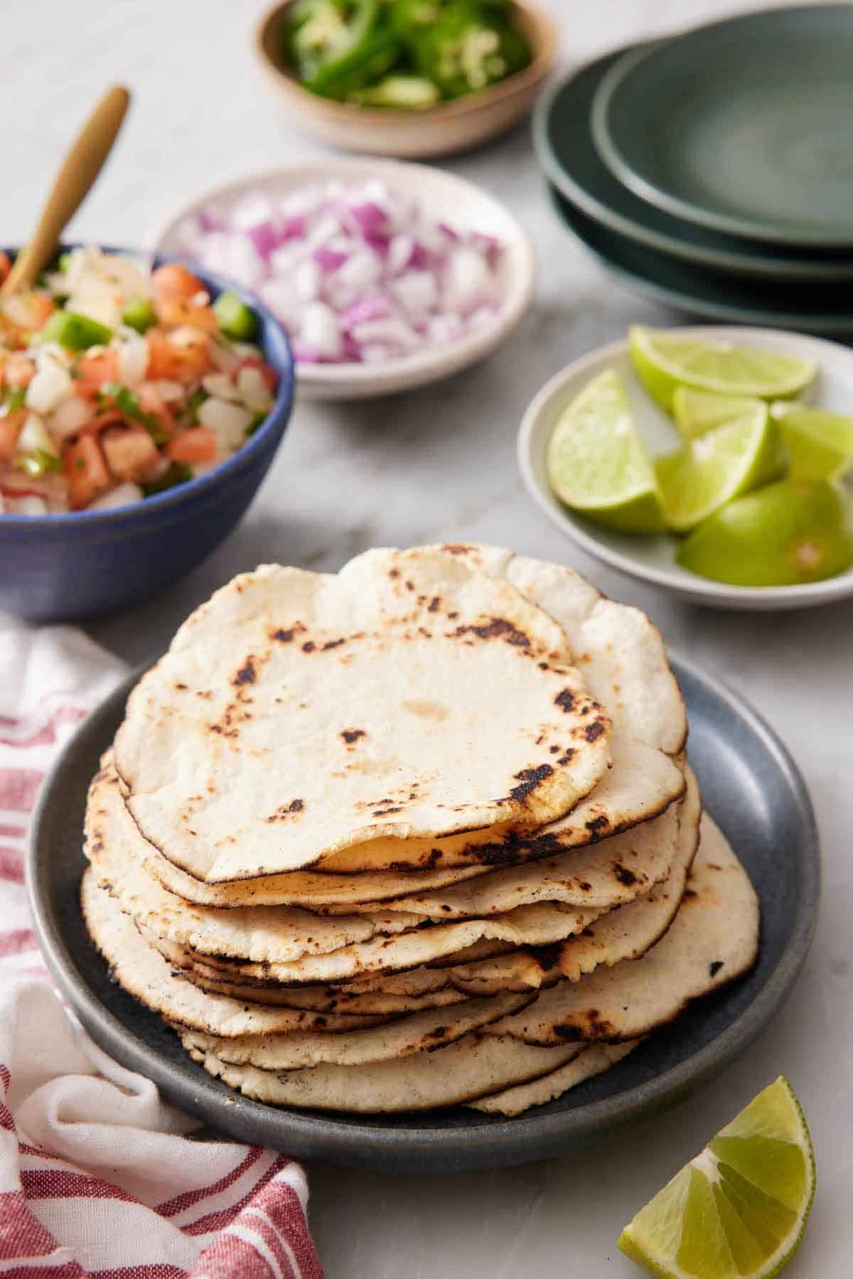 A stack of corn tortillas on a plate. A plate of limes, plate of diced onions, bowl of salsa, and bowl of jalapeno in the background along with a stack of plates.