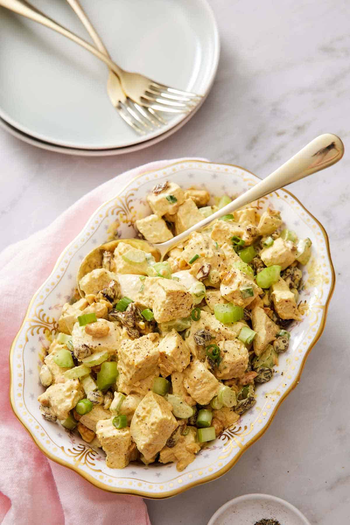 An overhead view of a platter of curried chicken salad with a spoon. A stack of plates and forks off to the side.